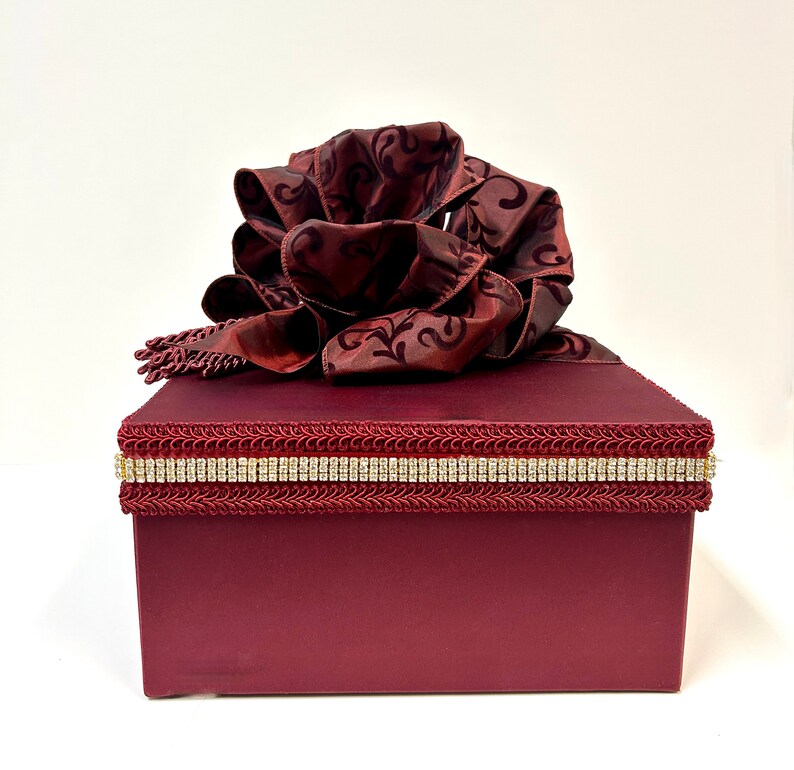 May include: A burgundy gift box with a large, ornate bow on top. The box is decorated with a rhinestone band and a decorative trim. The bow is made of a patterned fabric.