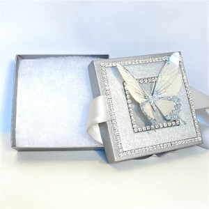 May include: A silver gift box with a white butterfly design and a white satin ribbon. The box is lined with white tissue paper.