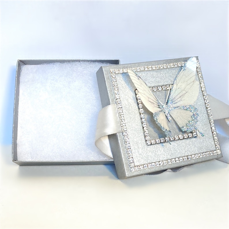 May include: A silver gift box with a white butterfly design and a white satin ribbon. The box is lined with white tissue paper.