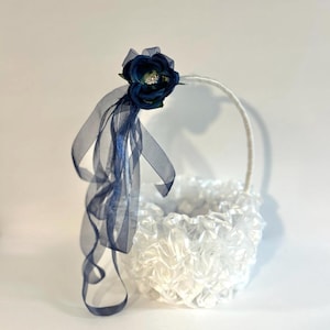 May include: A white flower girl basket with a ruffled texture. The basket is decorated with a navy blue flower and sheer ribbons. The handle is white and curved.