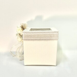 May include: White square box with a decorative lace trim around the top. The box has a small window on the front. A white flower bouquet is next to the box.