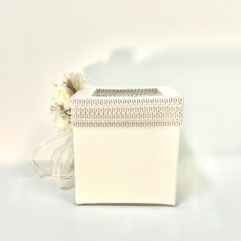 May include: White square box with a decorative lace trim around the top. The box has a small window on the front. A white flower bouquet is next to the box.