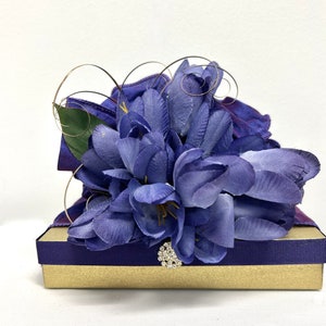 May include: A purple gift box with a satin ribbon and a bouquet of purple silk flowers. A sparkly rhinestone embellishment is attached to the ribbon.