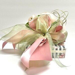 K&ouml;nnte beinhalten: Eine Geschenkbox, verpackt mit rosa und gr&uuml;nen transparenten B&auml;ndern und einer gro&szlig;en Schleife. Die Schleife ist mit einer glitzernden Verzierung versehen. Die Box ist mit einem Blumen- und Streifenmuster verziert.