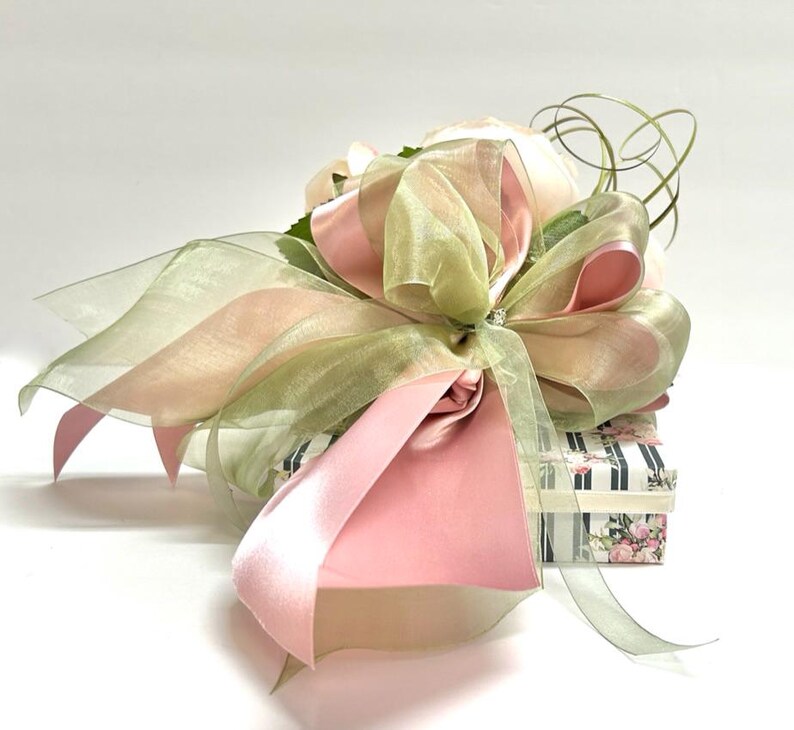 K&ouml;nnte beinhalten: Eine Geschenkbox, verpackt mit rosa und gr&uuml;nen transparenten B&auml;ndern und einer gro&szlig;en Schleife. Die Schleife ist mit einer glitzernden Verzierung versehen. Die Box ist mit einem Blumen- und Streifenmuster verziert.