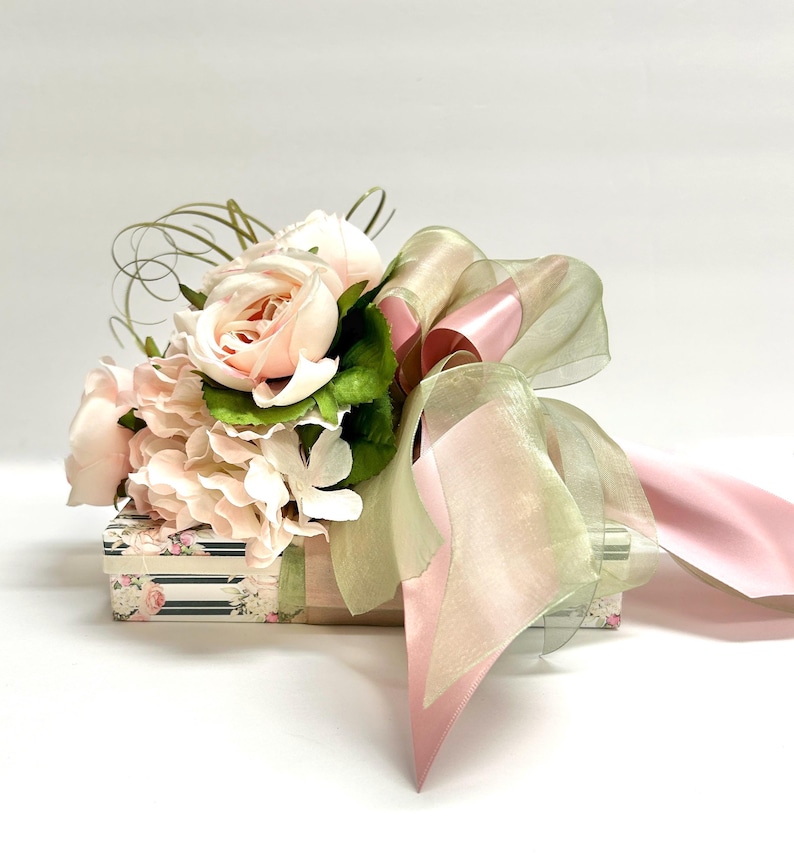 K&ouml;nnte beinhalten: Eine dekorative Geschenkbox mit einem Bouquet aus hellrosa Rosen, Hortensien und Gr&uuml;n. Die Box ist mit einem durchsichtigen, hellgr&uuml;nen und rosa Band umwickelt, was eine elegante Pr&auml;sentation ergibt. Die Box hat ein Blumen- und Streifenmuster.