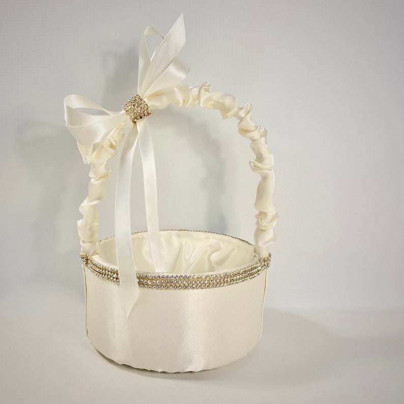 May include: A white satin flower girl basket with a ruffled handle and a large white satin bow with a rhinestone embellishment. The basket is decorated with a band of rhinestones around the top.