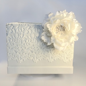 May include: A white tissue box cover with a detailed lace design on the front and sides. A large, white fabric flower with pearl and rhinestone embellishments decorates the top right. The top edge has a thin rhinestone trim.