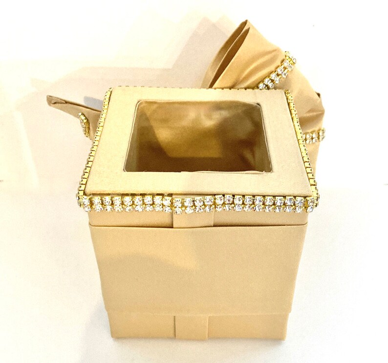 May include: A beige tissue box cover with a clear plastic window and a gold trim decorated with rhinestones.