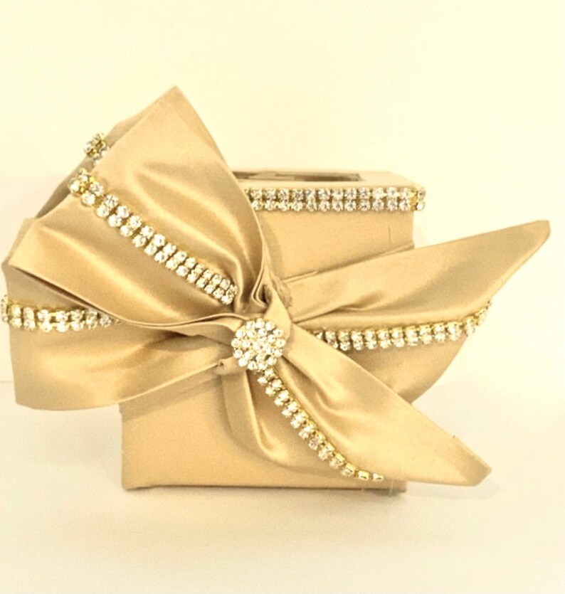 May include: A gold square box with a large satin bow and rhinestones. The bow is tied with a large rhinestone in the center.