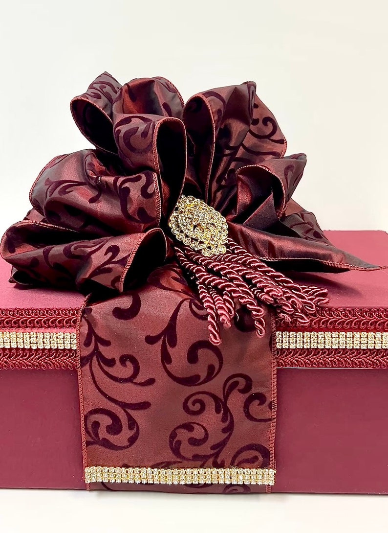 May include: A burgundy gift box with a large, ornate bow. The bow features a patterned design and is adorned with a jeweled brooch and tassel. The box is trimmed with rhinestone accents.