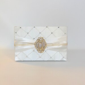 May include: A white satin box with a quilted pattern and a large, oval-shaped rhinestone brooch in the center. The box is tied with a white satin ribbon.