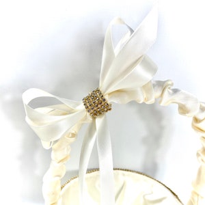 May include: A white satin ring bearer pillow with a gold and crystal embellishment in the centre. The pillow is tied with a large white satin bow.