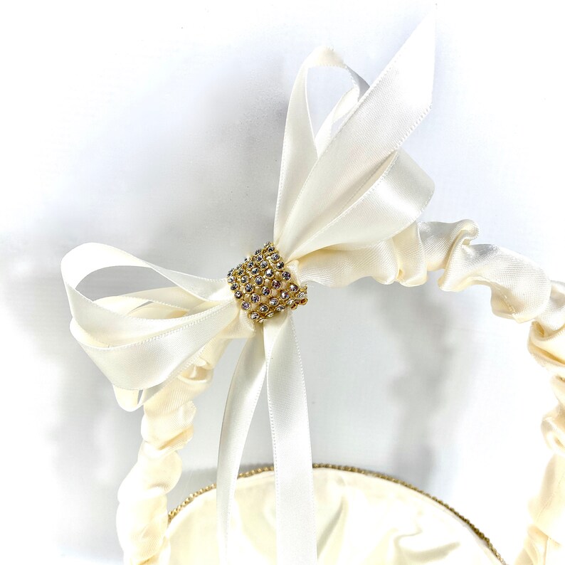 May include: A white satin ring bearer pillow with a gold and crystal embellishment in the centre. The pillow is tied with a large white satin bow.