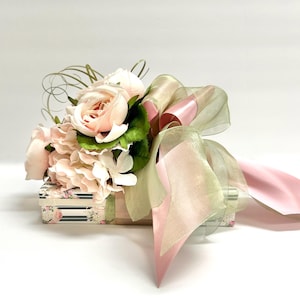 K&ouml;nnte beinhalten: Eine dekorative Geschenkbox, verziert mit einem Bouquet aus hellrosa Rosen, Hortensien und Gr&uuml;n. Die Box ist mit einem durchsichtigen, hellgr&uuml;nen und rosa Band umwickelt, was eine zarte und elegante Pr&auml;sentation ergibt. Die Box hat ein Blumen- und Streifenmuster.
