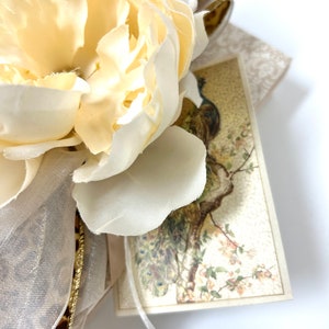 Puede incluir: Una flor de seda blanca con una cinta dorada y una tarjeta vintage con una ilustraci&oacute;n de un pavo real.