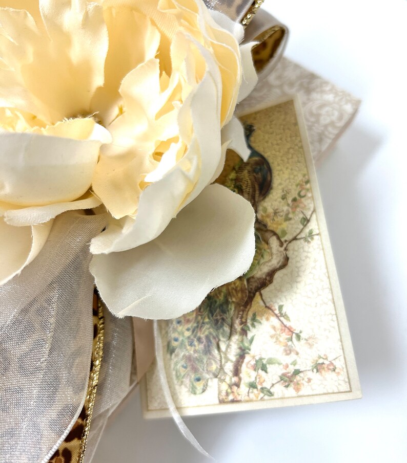 Puede incluir: Una flor de seda blanca con una cinta dorada y una tarjeta vintage con una ilustraci&oacute;n de un pavo real.
