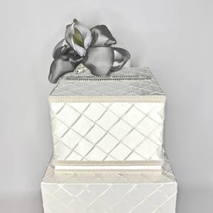 May include: White wedding card box with a diamond pattern and a silver ribbon bow on top. The box is two tiers high.