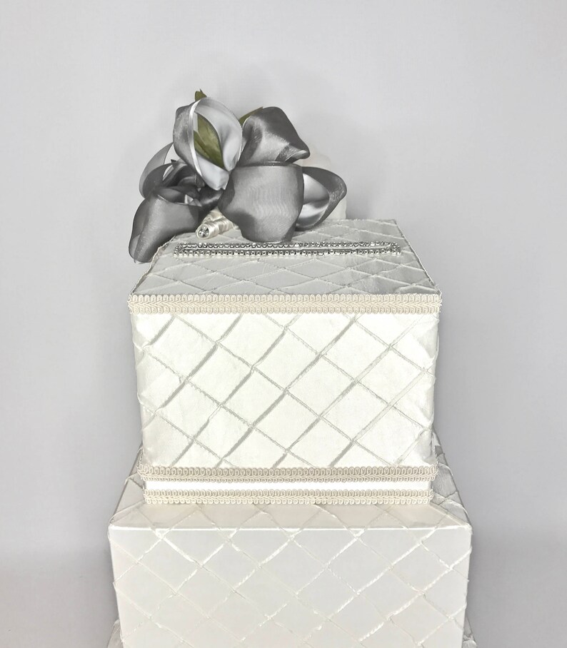 May include: White wedding card box with a diamond pattern and a silver ribbon bow on top. The box is two tiers high.