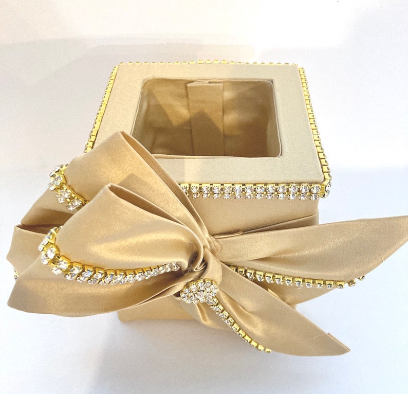 May include: A square-shaped tissue box cover with a gold satin finish and a clear window. The cover is decorated with a large gold satin bow and a border of rhinestones.