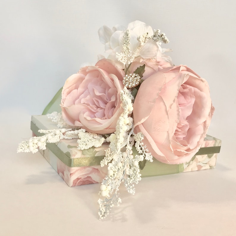 K&ouml;nnte beinhalten: Eine hellgr&uuml;ne Geschenkbox mit Blumenmuster ist mit einem Strau&szlig; rosa Seidenrosen und wei&szlig;en Zweigen verziert. Die Rosen haben eine zarte, gekr&auml;uselte Textur.
