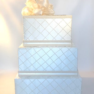 May include: A three-tiered white gift box with a diamond pattern and a white satin flower on top. The box is perfect for a wedding or other special occasion.