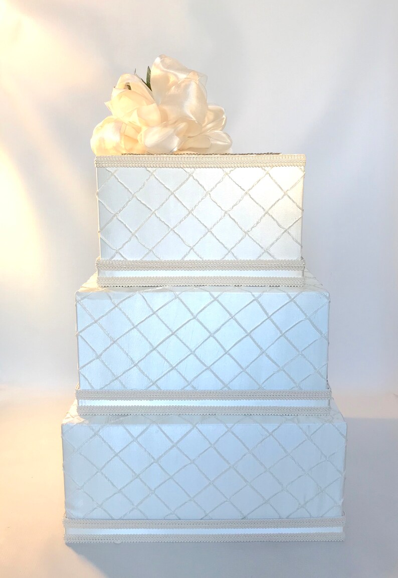 May include: A three-tiered white gift box with a diamond pattern and a white satin flower on top. The box is perfect for a wedding or other special occasion.