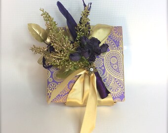 Puede incluir: Una pequeña caja de regalo morada y dorada con una cinta dorada y un arreglo floral morado y dorado con un broche de pedrería.