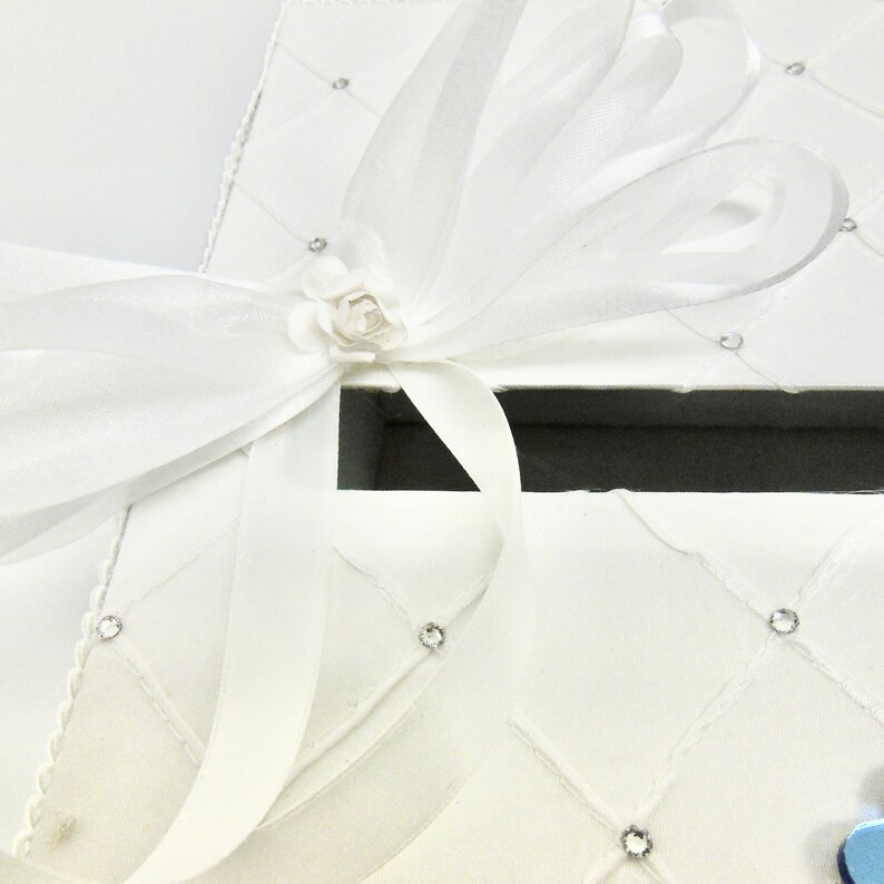 May include: White gift box with a white satin ribbon and a white flower. The box is decorated with small rhinestones.