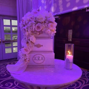 May include: A three-tiered white cake decorated with a cascade of white and pink roses. The cake has gold accents and the initials "BRM". A lit candle in a glass cylinder sits on a round table.