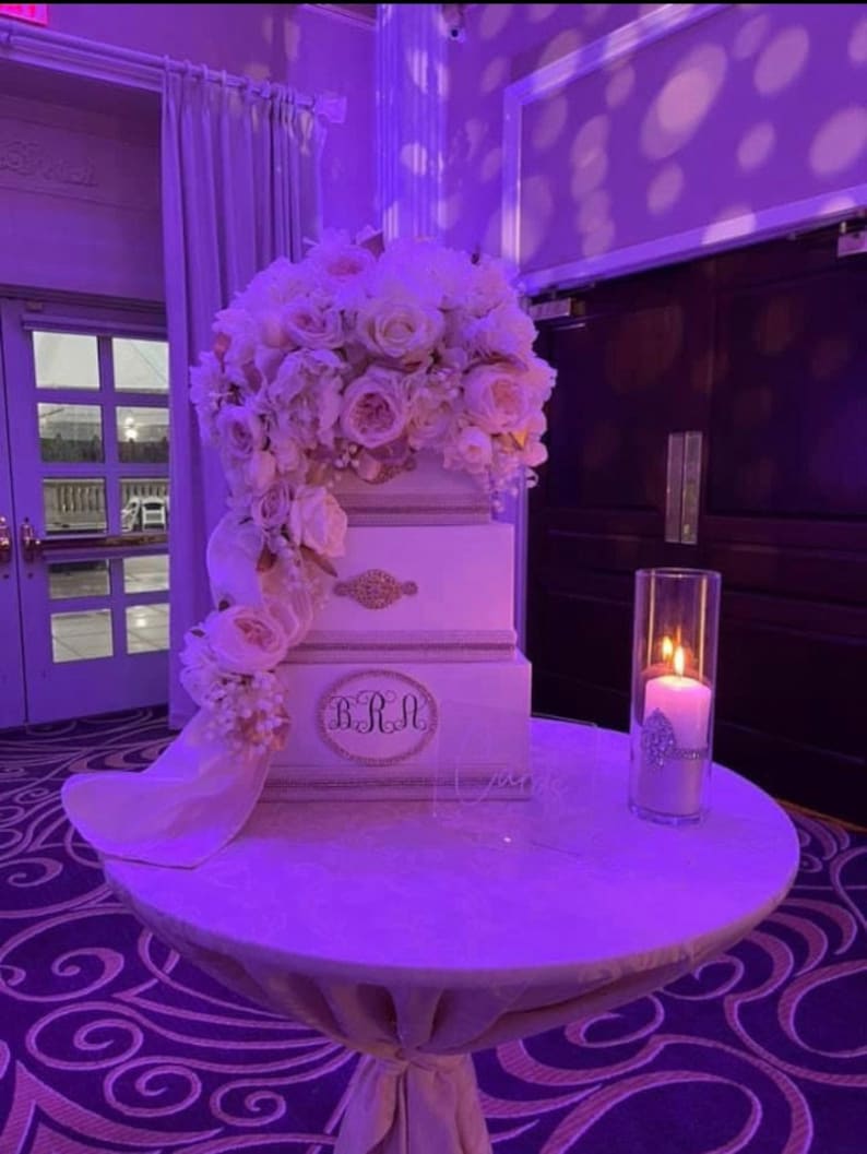 May include: A three-tiered white cake decorated with a cascade of white and pink roses. The cake has gold accents and the initials "BRM". A lit candle in a glass cylinder sits on a round table.