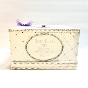 May include: A white gift box with a quilted design and rhinestones. The box has a pink and silver oval label with the text "God Bless Devyn Marseille Reed".