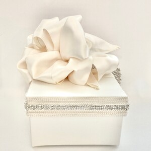 May include: Ivory-colored gift box with a large, flowing satin bow on top. The box is adorned with a decorative band of rhinestones and a textured trim. A stylish presentation for special occasions.