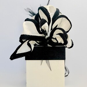 May include: A white gift box with a black ribbon and a large white bow with black trim. The bow is made of fabric and has a ruffled appearance.