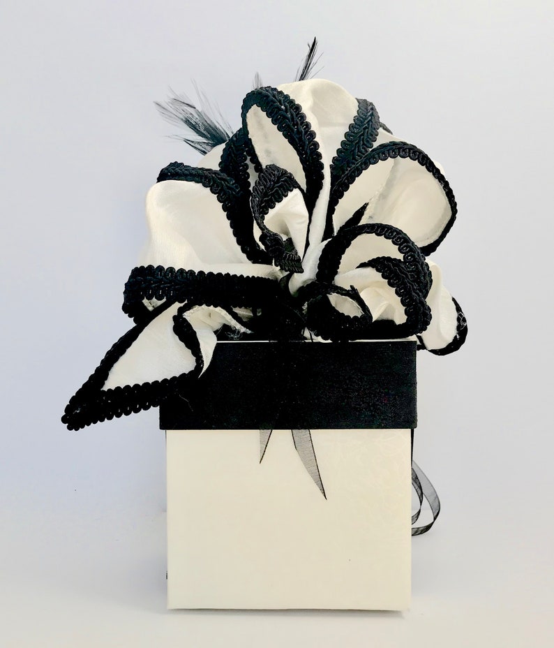 May include: A white gift box with a black ribbon and a large white bow with black trim. The bow is made of fabric and has a ruffled appearance.