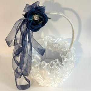 May include: A white flower girl basket with a ruffled design. It features a navy blue flower and sheer navy ribbons. The handle is white. The basket is set against a white background.