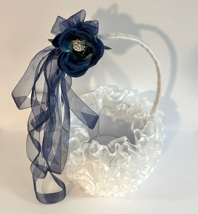 May include: A white flower girl basket with a ruffled design. It features a navy blue flower and sheer navy ribbons. The handle is white. The basket is set against a white background.