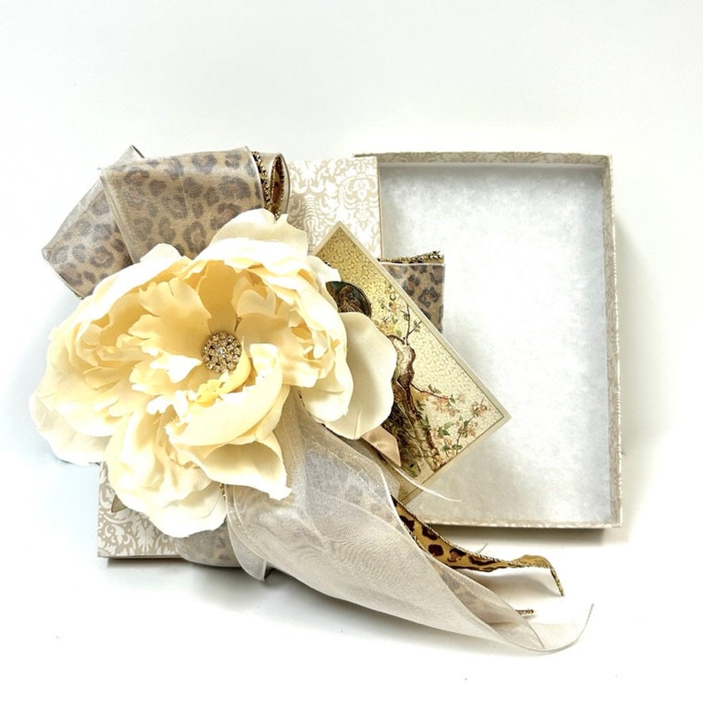 Puede incluir: Una caja de regalo blanca con una cinta de estampado de leopardo beige y marr&oacute;n y una gran flor blanca con un centro de pedrer&iacute;a. La caja est&aacute; abierta y el interior est&aacute; forrado con papel de seda blanco.