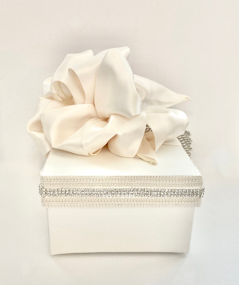 May include: Ivory-colored gift box with a large, flowing satin bow on top. The box is adorned with a decorative band of rhinestones and a textured trim. A stylish presentation for special occasions.