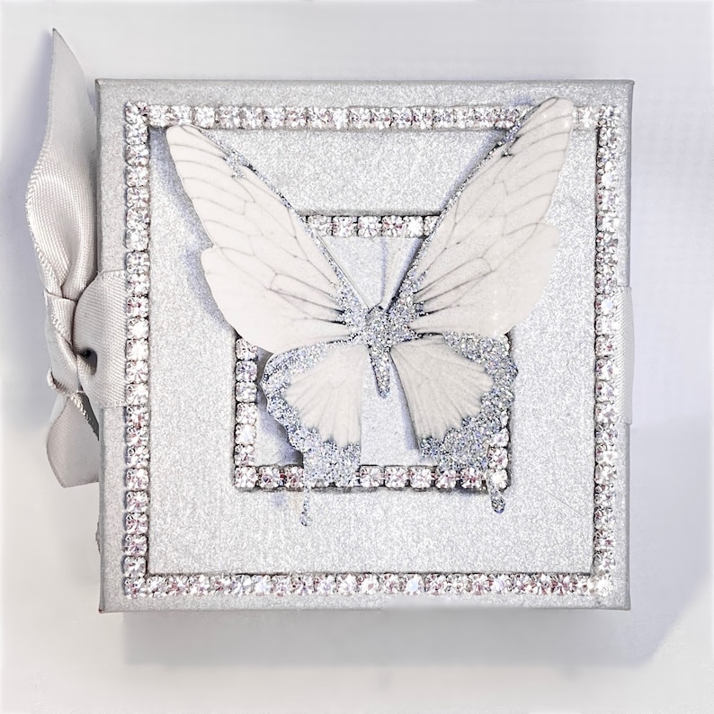 May include: A silver glitter box with a white butterfly design. The box is decorated with a silver rhinestone border.