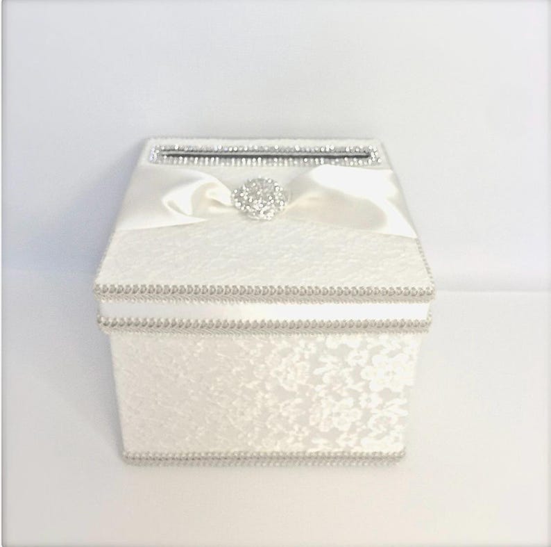 K&ouml;nnte beinhalten: Wei&szlig;e Hochzeitskartenbox mit einem Satinband und einem Strassstein-Verzierungen. Die Box hat ein Spitzenmuster und einen Schlitz f&uuml;r Karten.