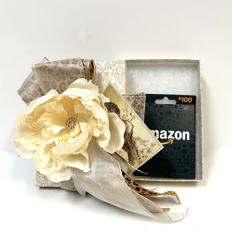 Puede incluir: Una caja de regalo con una flor blanca y una tarjeta de regalo de Amazon de 100 &euro;. La caja est&aacute; envuelta en papel estampado beige y marr&oacute;n con una cinta beige.