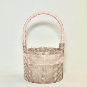 May include: A sparkling, blush-toned handbag with a rounded base and a soft pink handle. The body of the bag is covered in small, shimmering embellishments, with a horizontal band of lighter pink. The handle is a soft, velvety pink.