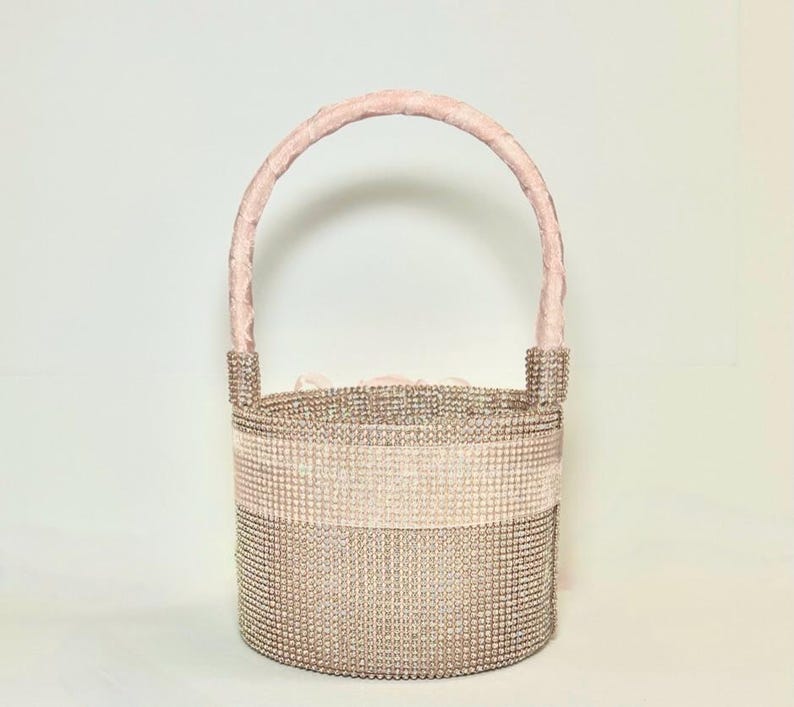 May include: A sparkling, blush-toned handbag with a rounded base and a soft pink handle. The body of the bag is covered in small, shimmering embellishments, with a horizontal band of lighter pink. The handle is a soft, velvety pink.