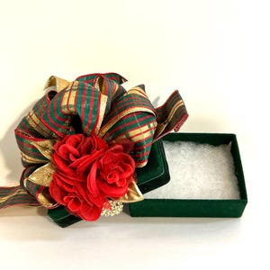 May include: A green velvet gift box with a red and gold striped ribbon and a red fabric flower on top. The box is open and the inside is lined with white tissue paper.
