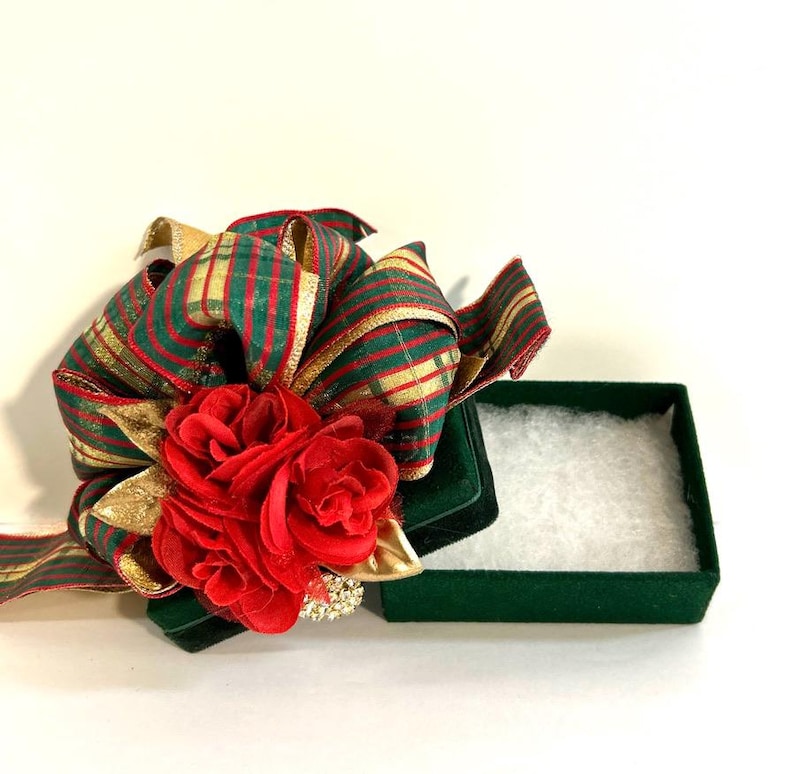 May include: A green velvet gift box with a red and gold striped ribbon and a red fabric flower on top. The box is open and the inside is lined with white tissue paper.