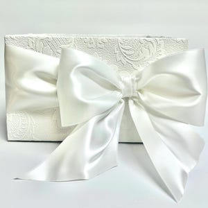 May include: A rectangular white gift box with a large, satin bow. The box has a textured, embossed floral pattern. The bow is made of a shiny, white satin material. The box is suitable for weddings, birthdays, or other special occasions.