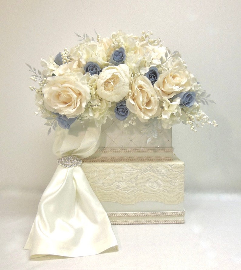 Pu&ograve; includere: Una scatola per biglietti di nozze bianca a due livelli con un nastro di raso e un abbellimento di strass. Il livello superiore &egrave; decorato con un arrangiamento floreale bianco e blu.