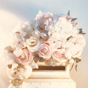 May include: A white card box adorned with a large arrangement of cream and blush-colored roses, peonies, and hydrangeas. The box features a decorative ribbon, a rhinestone brooch, and a slot for cards. The overall theme is wedding or event decor.