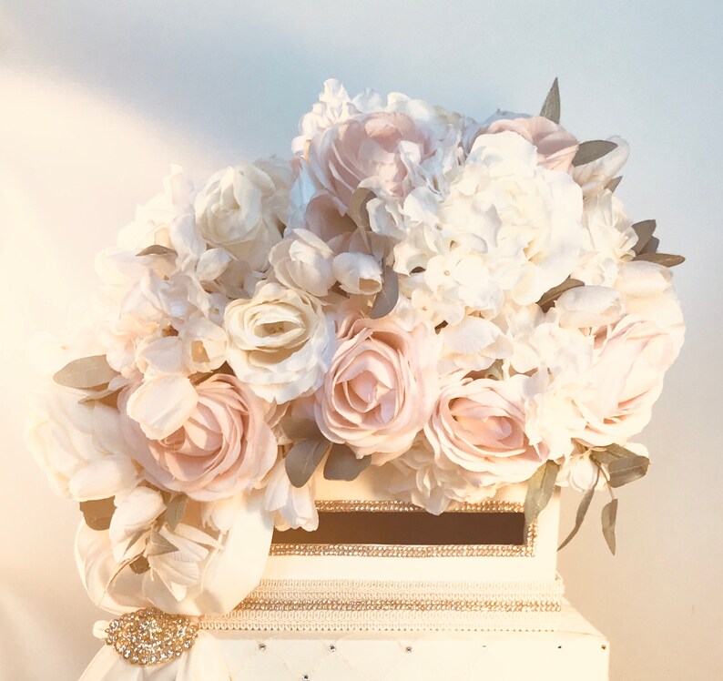 May include: A white card box adorned with a large arrangement of cream and blush-colored roses, peonies, and hydrangeas. The box features a decorative ribbon, a rhinestone brooch, and a slot for cards. The overall theme is wedding or event decor.